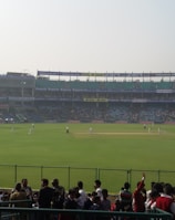 A cricket stadium filled with spectators, with players on the field wearing white uniforms. The stands are populated, and large floodlights are visible. Advertisements are displayed around the stadium, and a bright digital screen is on the left side.