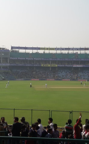 A cricket stadium filled with spectators, with players on the field wearing white uniforms. The stands are populated, and large floodlights are visible. Advertisements are displayed around the stadium, and a bright digital screen is on the left side.