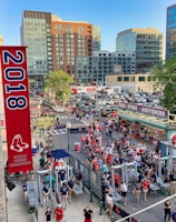 A vibrant outdoor scene featuring a large crowd of people gathered near a sports venue. A prominent red banner with '2018' and a sports logo hangs from a brick building. Nearby, there is a sign for a team store and souvenir shop. Surrounding the event are several modern high-rise buildings and a parking lot full of cars. Tall trees are visible in the background.