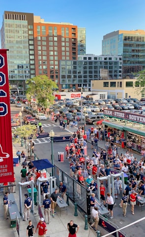 A vibrant outdoor scene featuring a large crowd of people gathered near a sports venue. A prominent red banner with '2018' and a sports logo hangs from a brick building. Nearby, there is a sign for a team store and souvenir shop. Surrounding the event are several modern high-rise buildings and a parking lot full of cars. Tall trees are visible in the background.