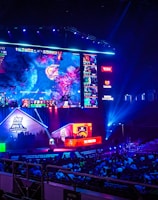 A large esports arena featuring a massive screen displaying a video game, surrounded by sponsor logos and team names. The auditorium is filled with spectators watching the gaming event. Neon blue and purple lights illuminate the stage, creating a vibrant atmosphere.