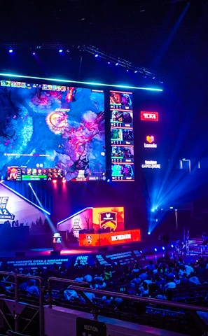 A large esports arena featuring a massive screen displaying a video game, surrounded by sponsor logos and team names. The auditorium is filled with spectators watching the gaming event. Neon blue and purple lights illuminate the stage, creating a vibrant atmosphere.