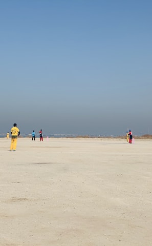 A group of people wearing colorful sports uniforms are playing cricket on a wide, open sandy ground. The sky is clear with a subtle gradient from blue to grey on the horizon. In the distance, there are structures that suggest a cityscape.