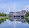 The scene features a large sports stadium with distinctive triangular white supports, located by a tranquil river. The stadium is surrounded by modern buildings and lush green trees. A bridge with multiple arches crosses over the river, and its reflection is visible on the calm water surface.