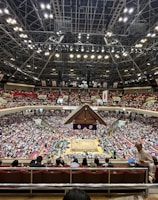A large indoor arena packed with spectators seated in tiers around a central platform. The platform features traditional Japanese decor, indicating it may be used for sumo wrestling. The atmosphere is bustling, with people filling the seats and many spectators visible in the foreground.