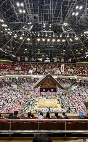 A large indoor arena packed with spectators seated in tiers around a central platform. The platform features traditional Japanese decor, indicating it may be used for sumo wrestling. The atmosphere is bustling, with people filling the seats and many spectators visible in the foreground.