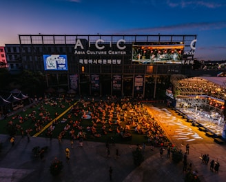An outdoor event at the Asia Culture Center features a large audience sitting on the grass, watching a live performance on a stage. The stage is brightly lit, contrasting with the darkening evening sky. A large screen displays the performers, and there's signage for the 'World Music Festival' on the buildings.
