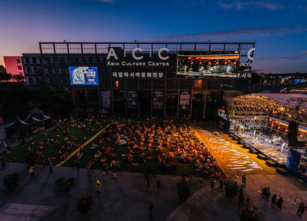 An outdoor event at the Asia Culture Center features a large audience sitting on the grass, watching a live performance on a stage. The stage is brightly lit, contrasting with the darkening evening sky. A large screen displays the performers, and there's signage for the 'World Music Festival' on the buildings.